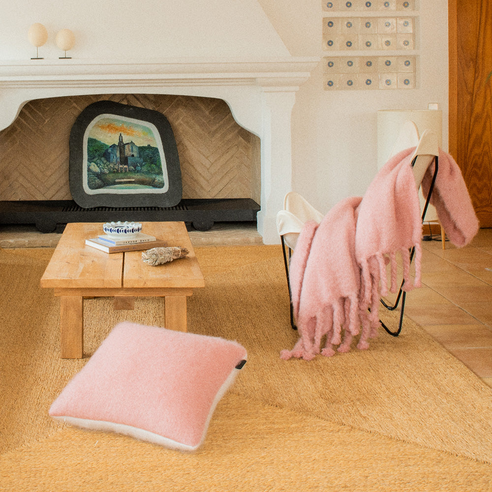 A living room with a fireplace, a table and a chair, where you’ll find a pillow and a pink mohair blanket.