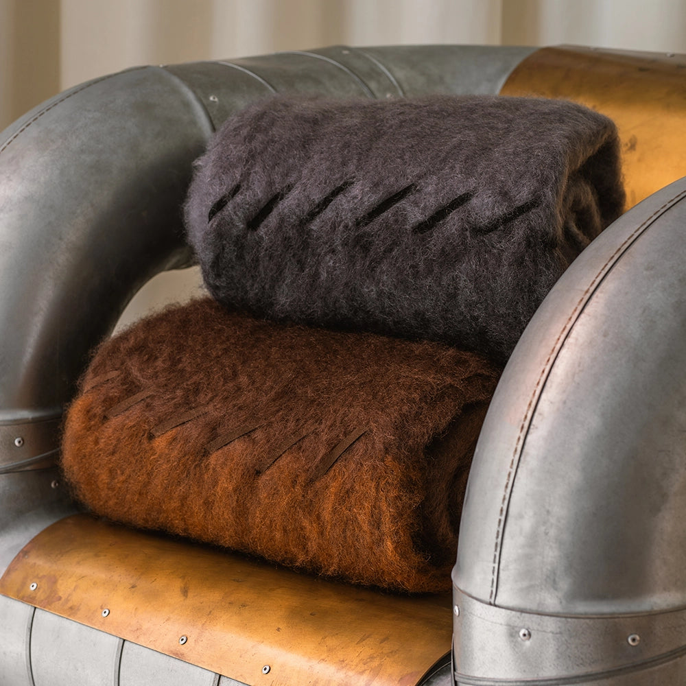 Two folded mohair blankets, one brown and one gray, on a vintage-style chair with metallic and leather accents.