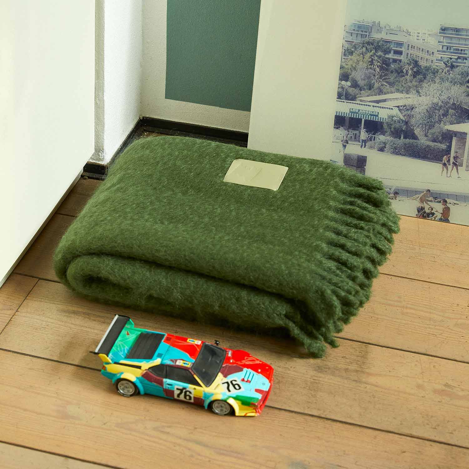 Inside a room, there is a green mohair blanket folded on the floor next to a toy car.