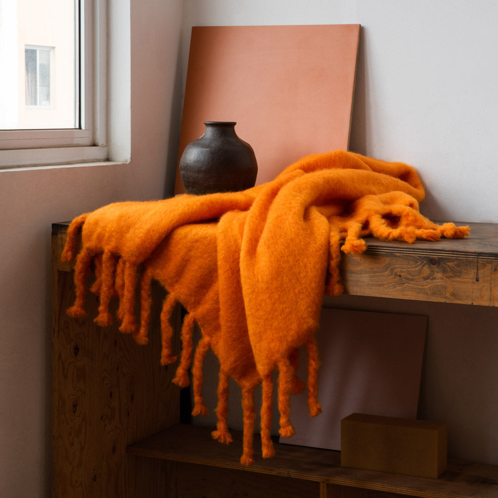 An orange mohair blanket on a wooden table, next to a clay vase and a window.