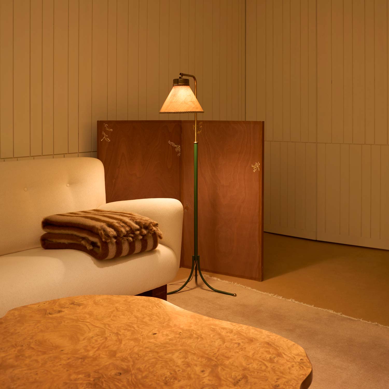 A living room with a lamp casting a warm glow over a wooden table and a white sofa, on which lies a striped mohair blanket in beige and brown.