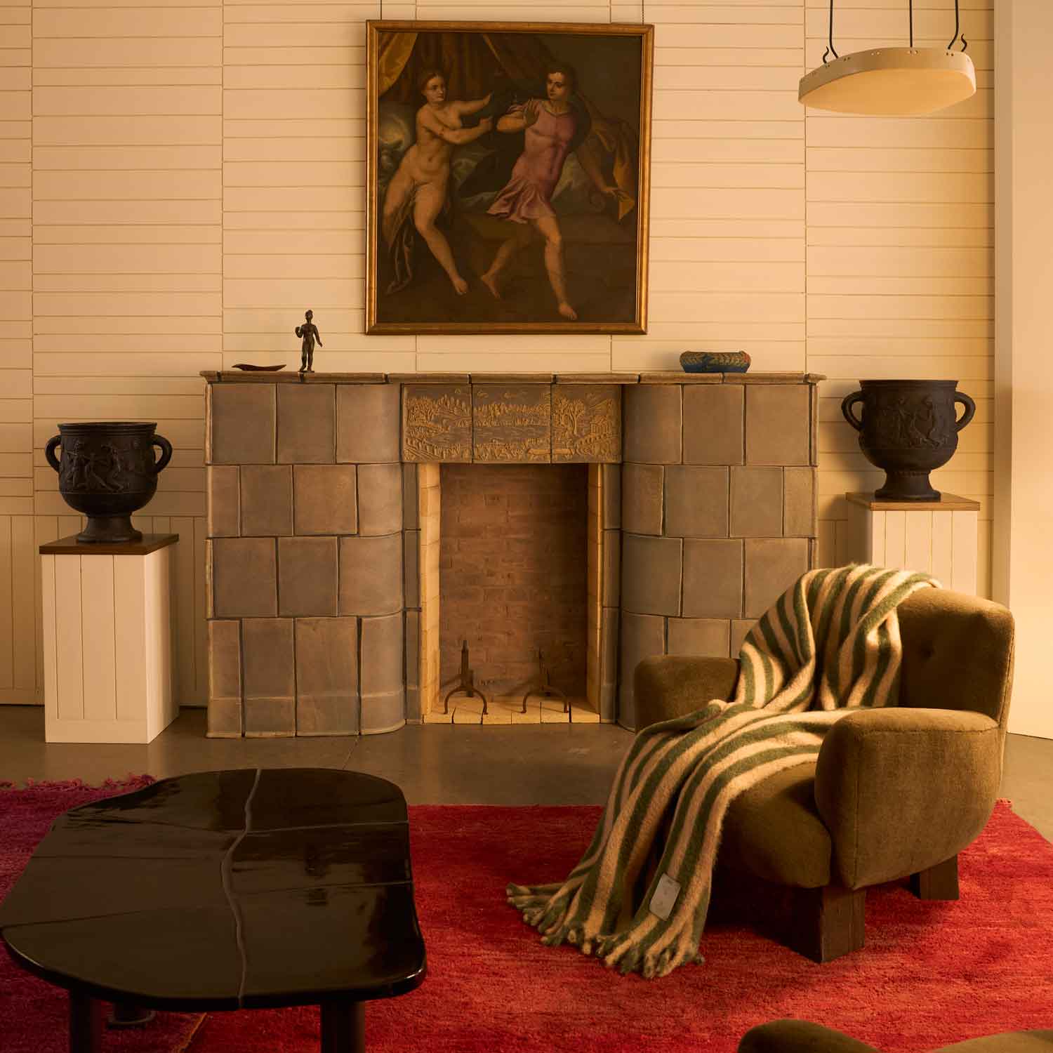 A living room with a fireplace in the centre. In front of it is a table and an armchair with a green and beige striped blanket draped over it. 