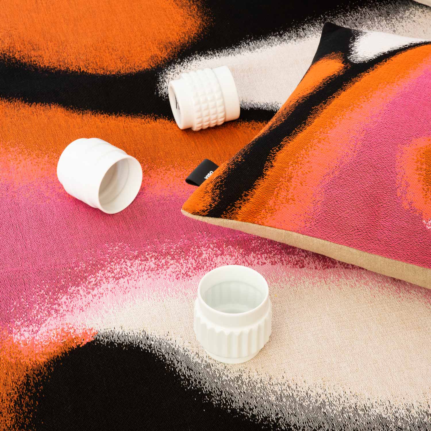 A close-up of a bed covered with a tapestry blanket in vibrant shades of pink and orange, with a pillow made from the same fabric and some white porcelain glasses placed on top.