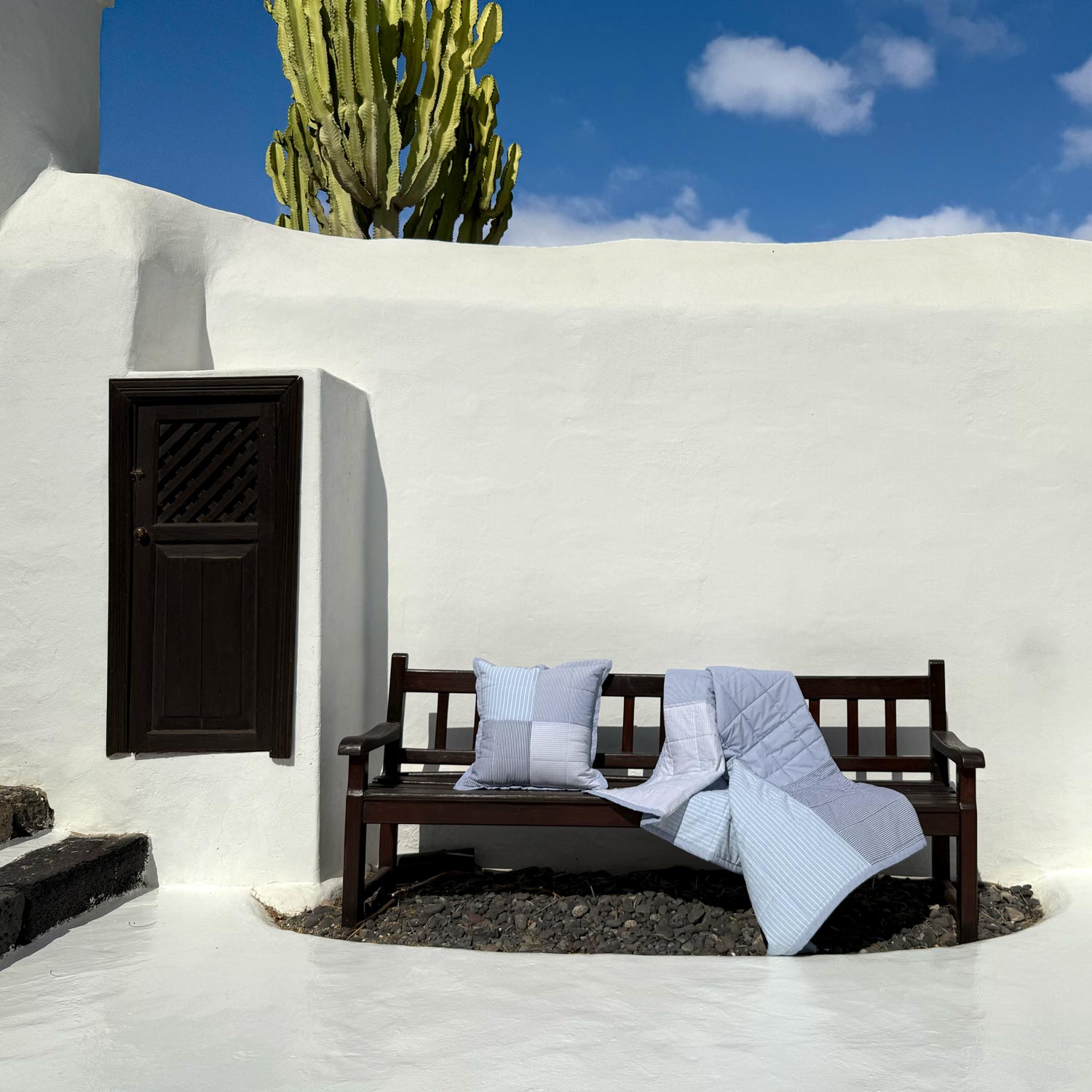 Wooden bench with pillow and blanket against a white wall and cactus in the background