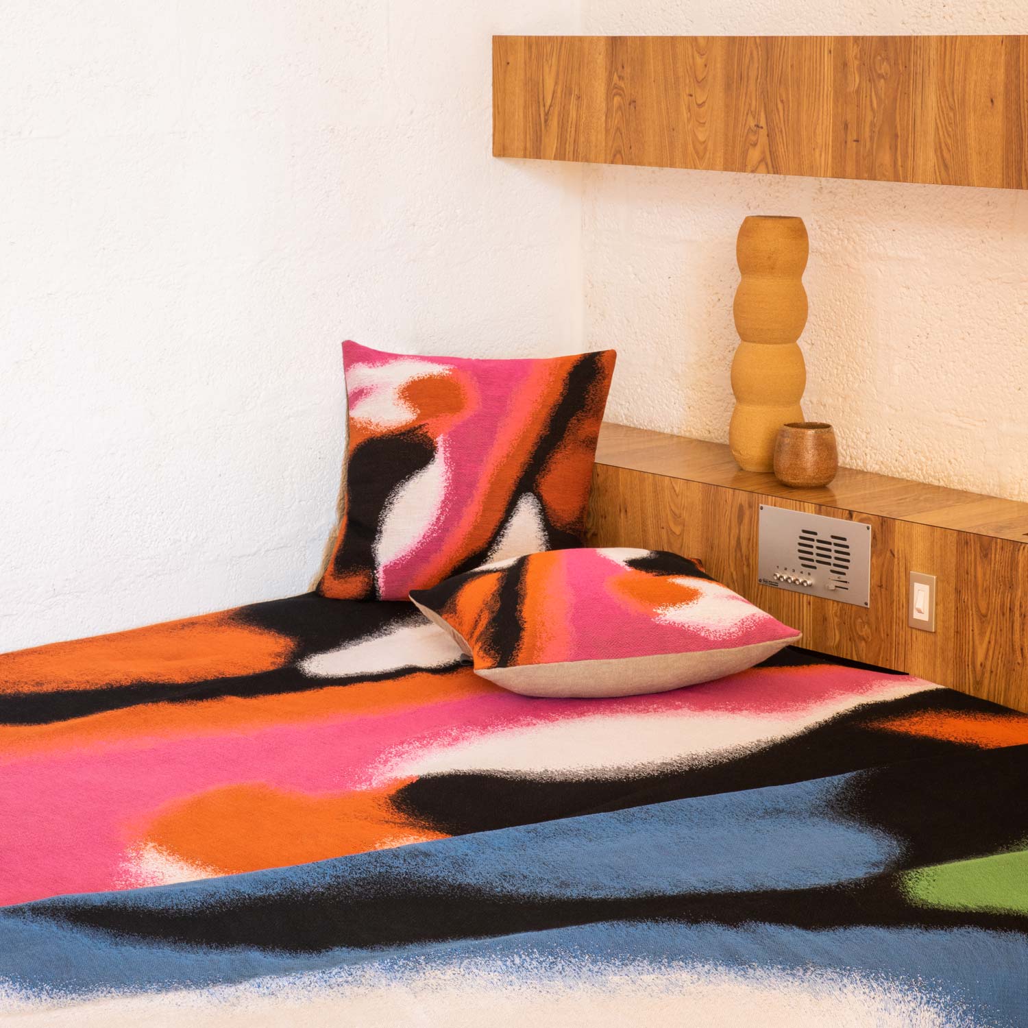 A close-up of a bed with blanket and tapestry pillows featuring an abstract design in bright colors and some ceramic vase behind.