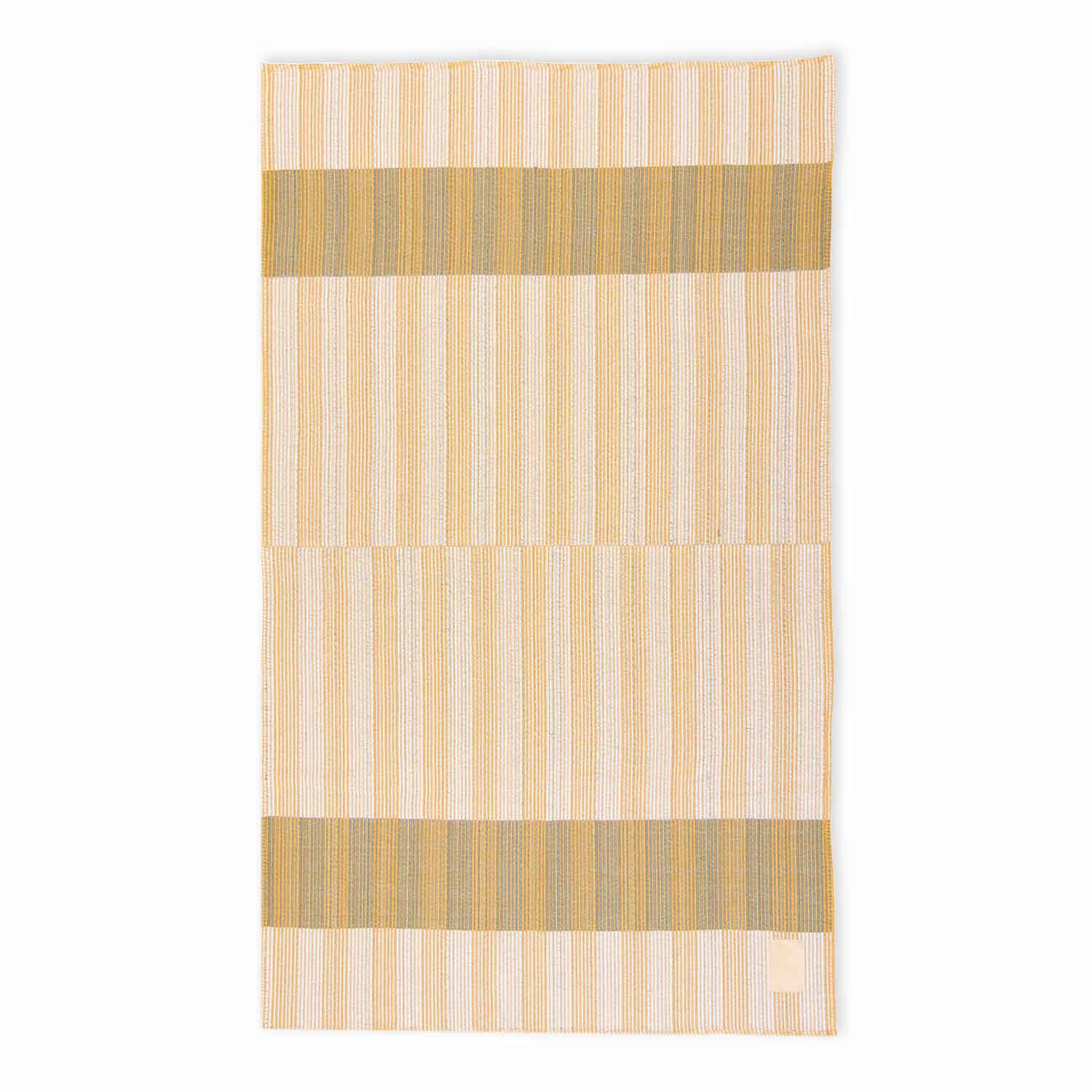 A flat striped cotton blanket in beige, tan, and gray tones, with a visible brand label on the bottom right.