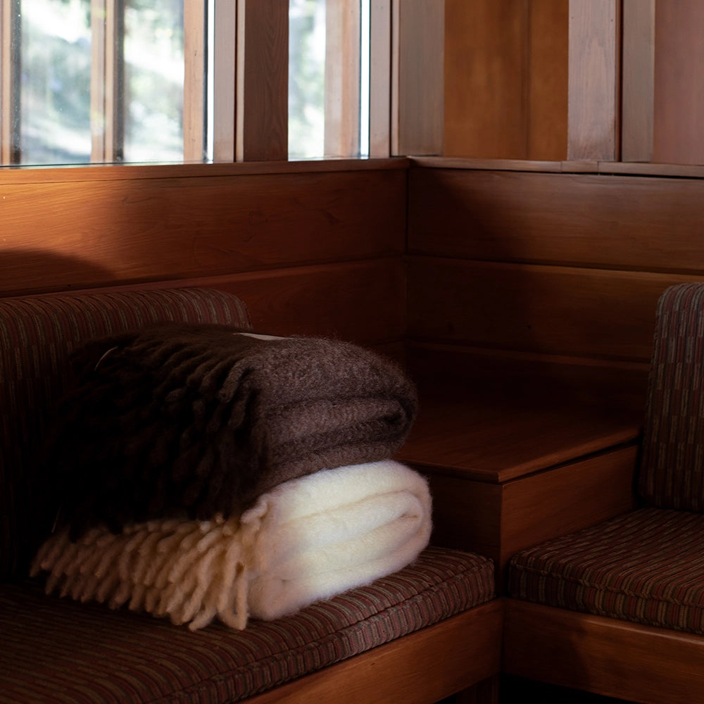 Stack of folded mohair blankets on a wooden bench with a window in the background – Ready to Hang X Viso Project