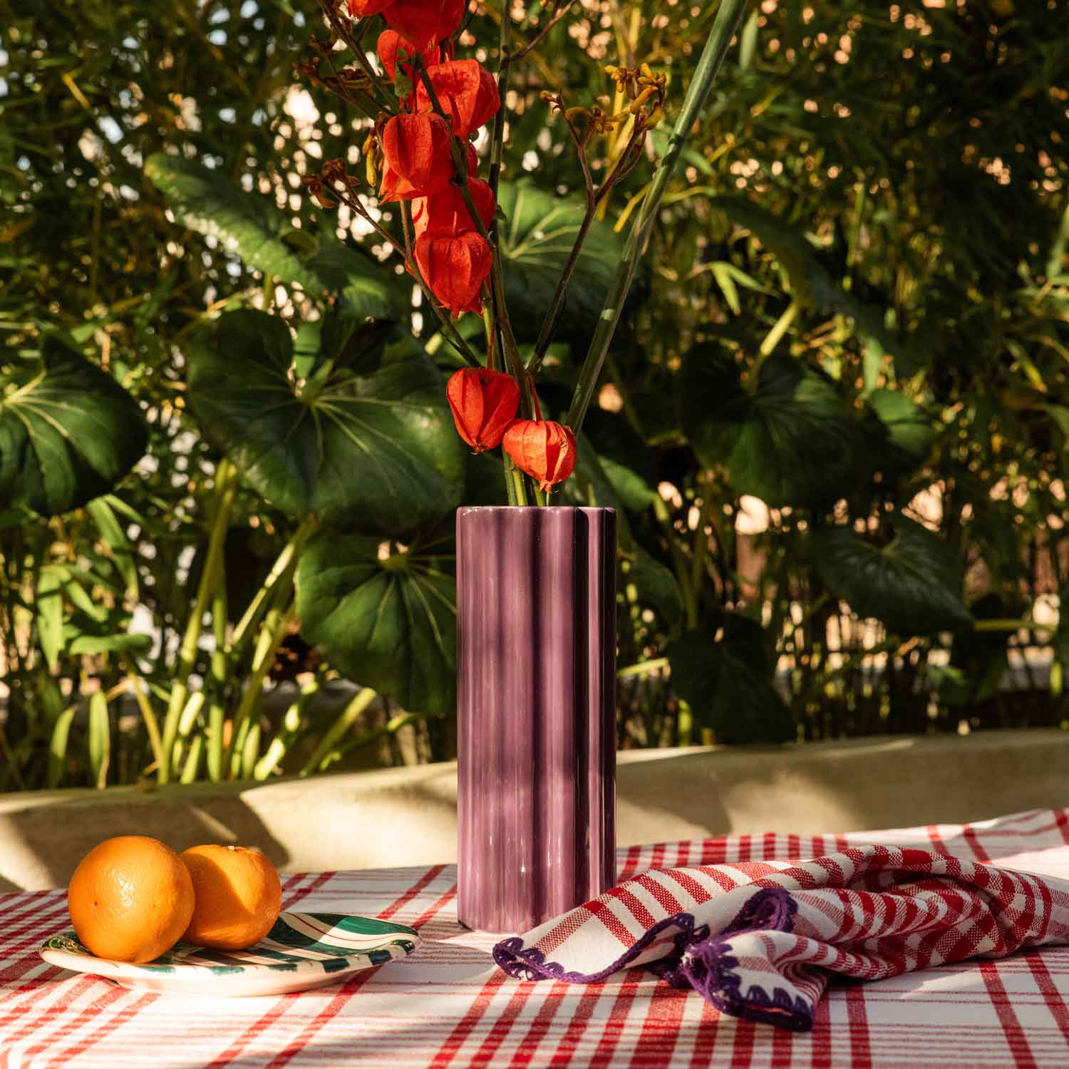 A table in a Mediterranean country house, laid with a tablecloth
and red and purple napkins, with ceramic crockery and a ceramic vase on top.