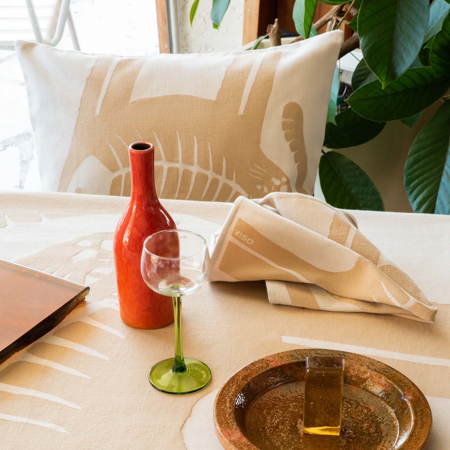 A table set for a meal, with a beige and cream napkin and tablecloth and a matching pillow on the chair featuring an illustration of some cats.