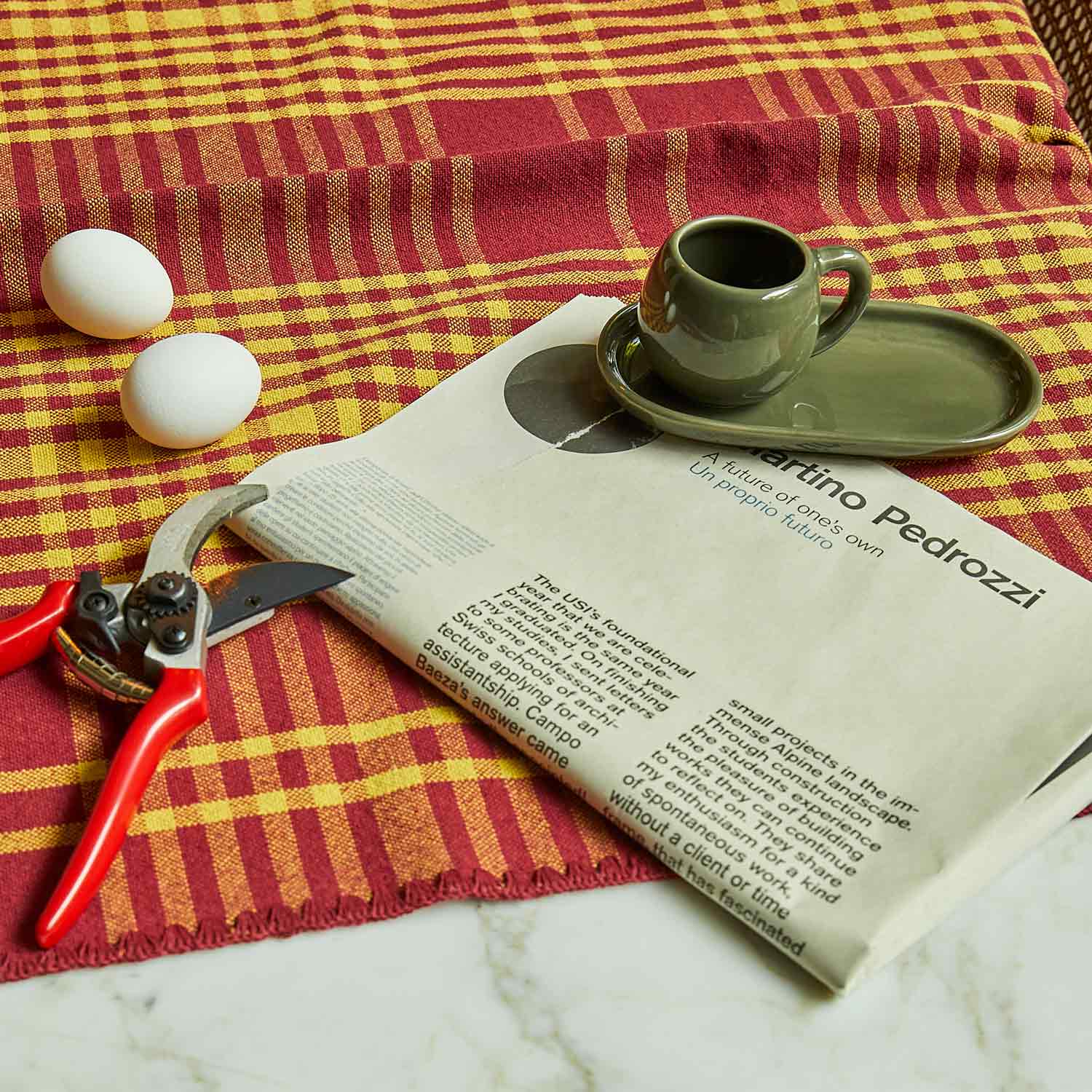 A close-up of a bordeaux and yellow chequered tablecloth, on which there is an egg, a branch-shaped carving knife and a green ceramic coffee cup.