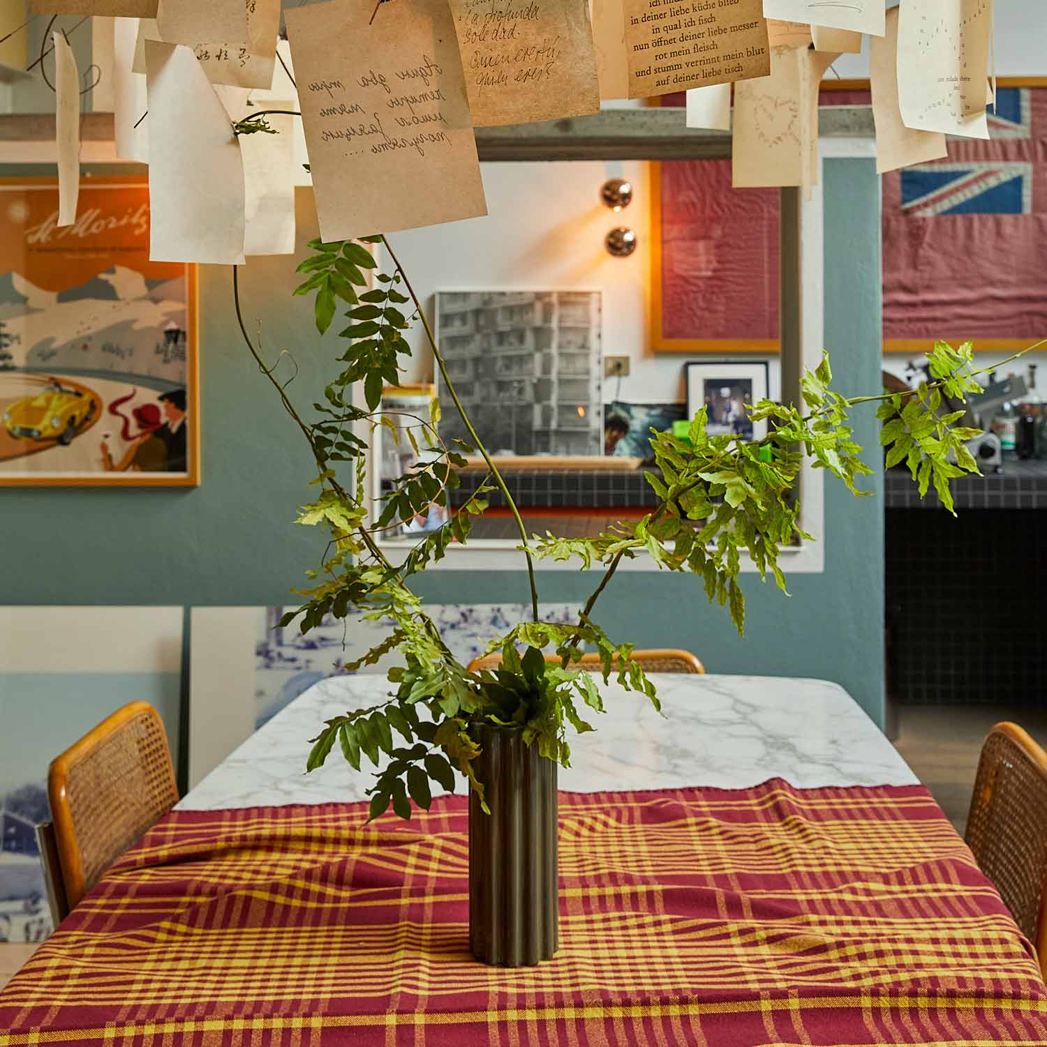 A living room with a lamp on a table covered with a burgundy and yellow chequered tablecloth and a green ceramic vase in the centre.