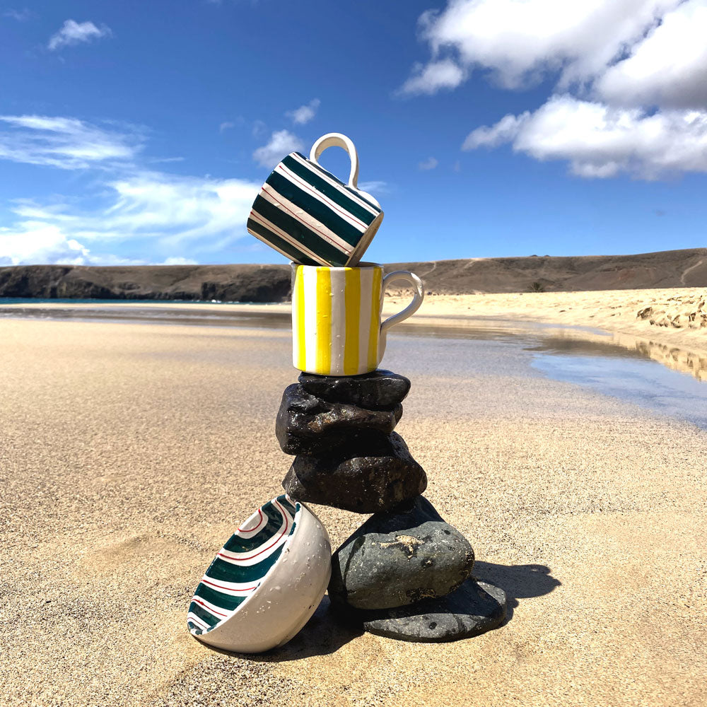 Mugs and bowls featuring a design of white, green and red stripes, and another with yellow and white stripes set against a backdrop of stones on the beach.