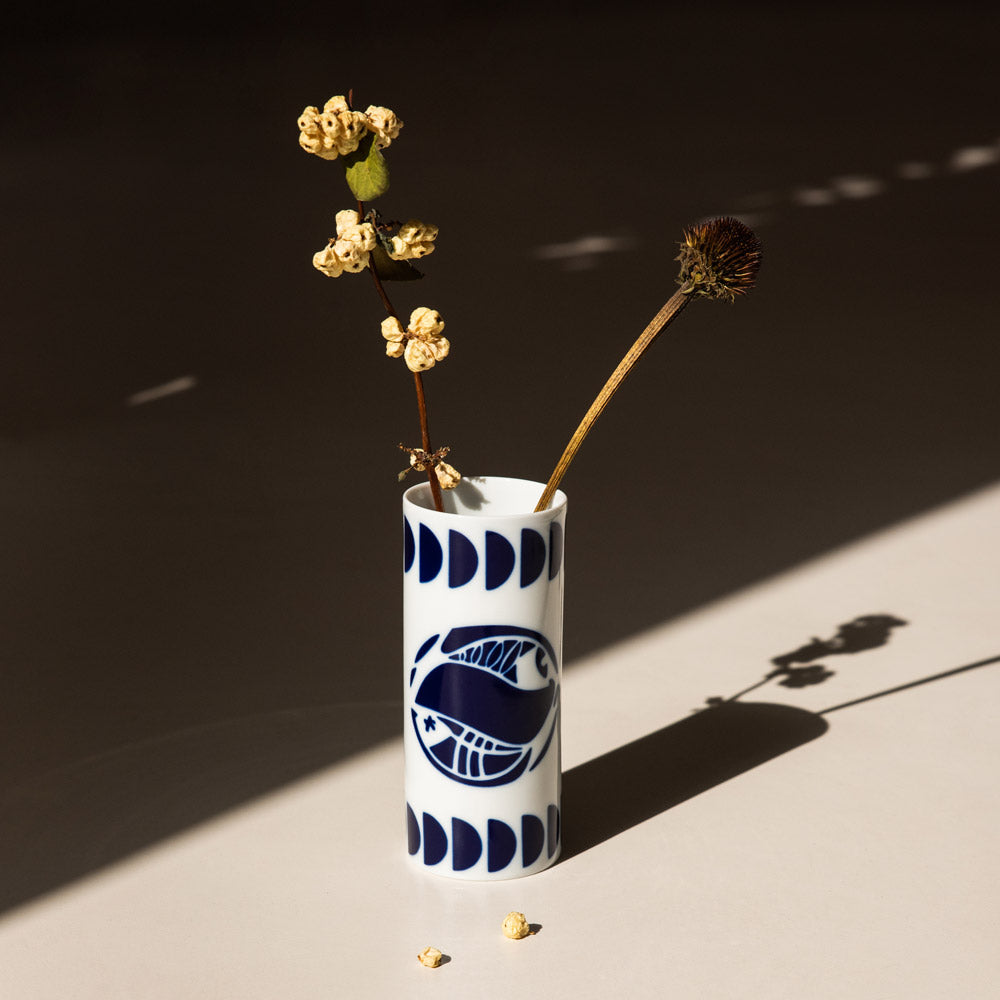 A mini white porcelain vase, painted in navy blue with each sign of the zodiac, featuring dried flowers and creating a play of shadows.