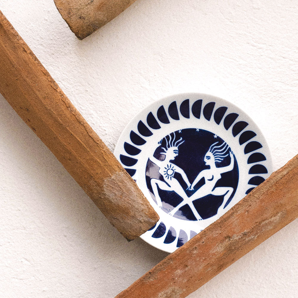 A small round white ceramic plate with a dark blue hand-painted folk art design of two figures as a gimini zodiac horoscope, styled on a textured white wall with rustic wooden props.