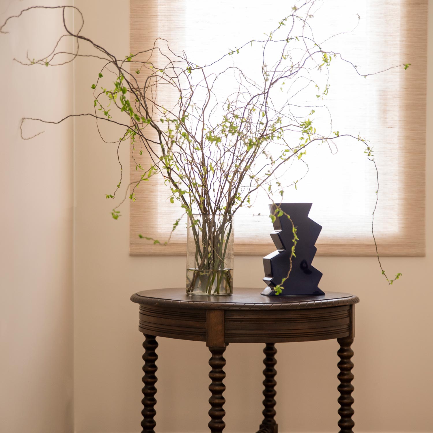 A navy-coloured, zigzag-shaped porcelain vase on a small hall table, next to a glass vase filled with plants.