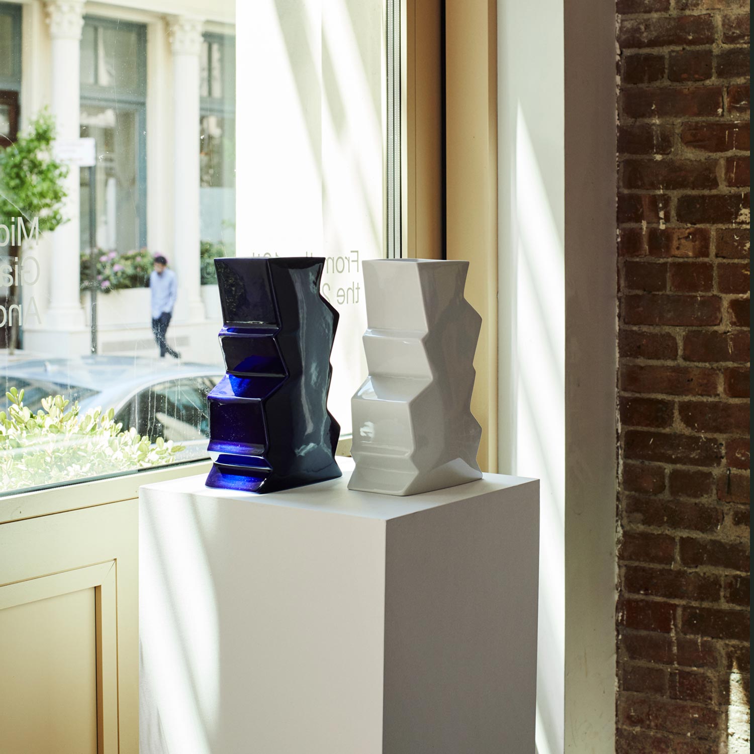 Two porcelain vases – one white and the other navy – on a pedestal in front of a window.
