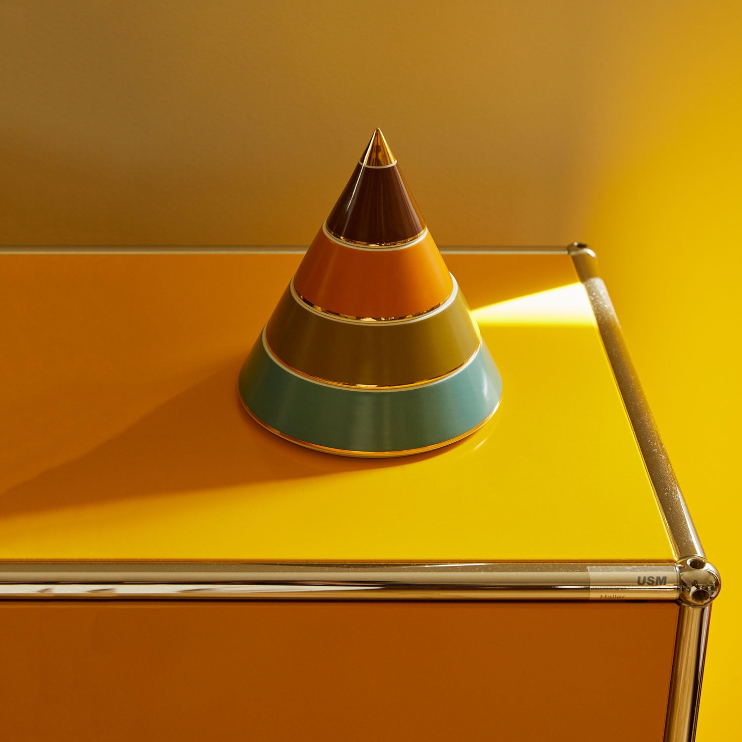 A multicoloured porcelain jewellery pyramid with gold-plated details, featuring layers of brown, green, blue and mustard on a yellow desk.