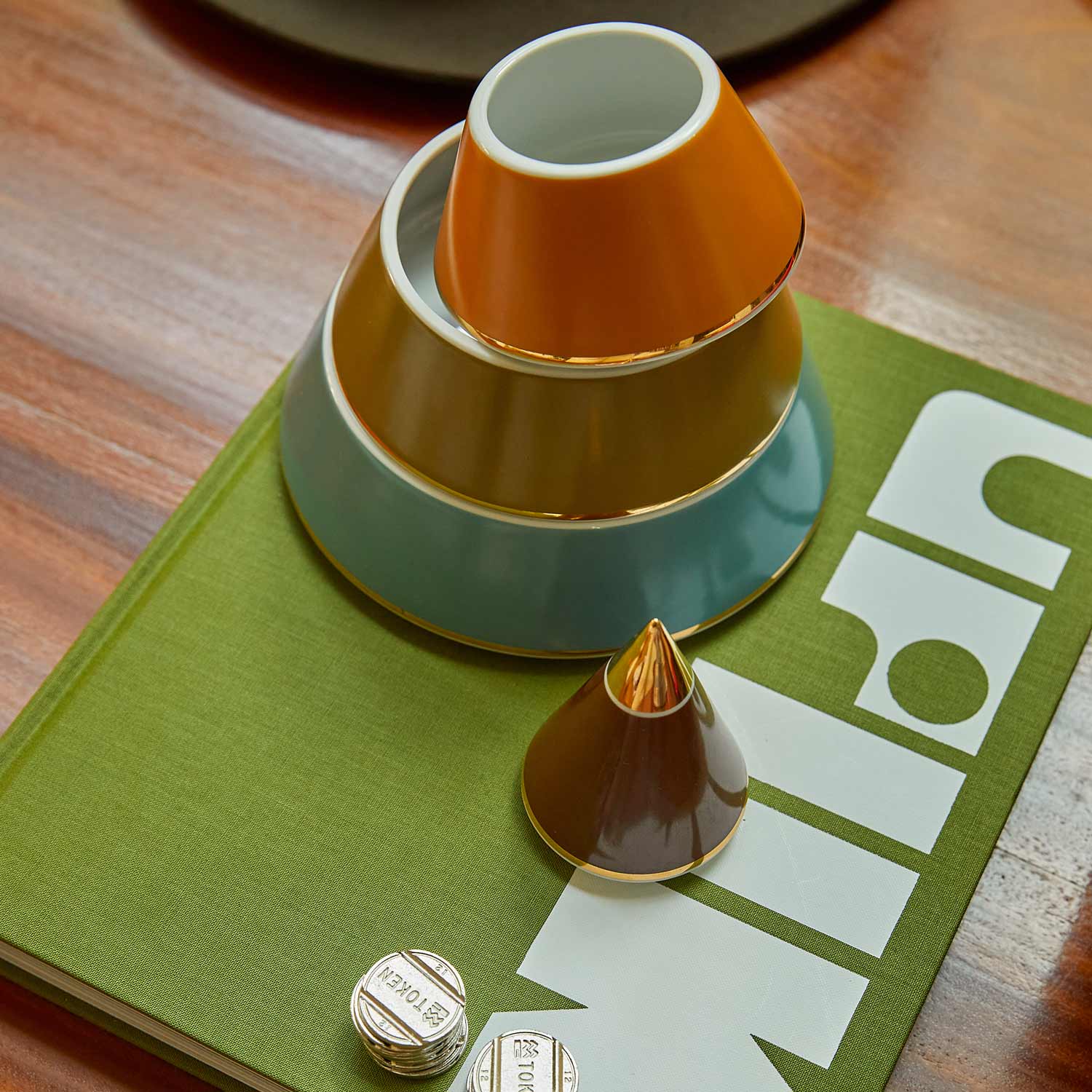 A multicoloured porcelain jewellery pyramid with gold-plated details, featuring layers of brown, green, blue and mustard on top of a green book and a table.