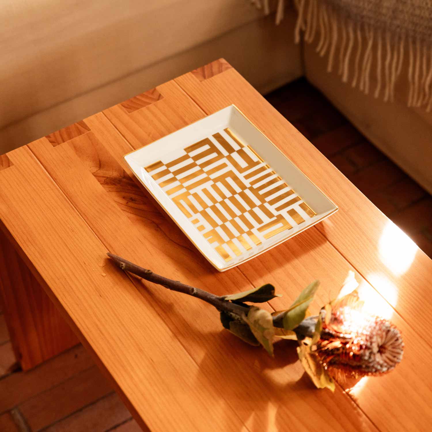 A white porcelain tray with an abstract gold pattern on a wooden table, next to some dry branches.