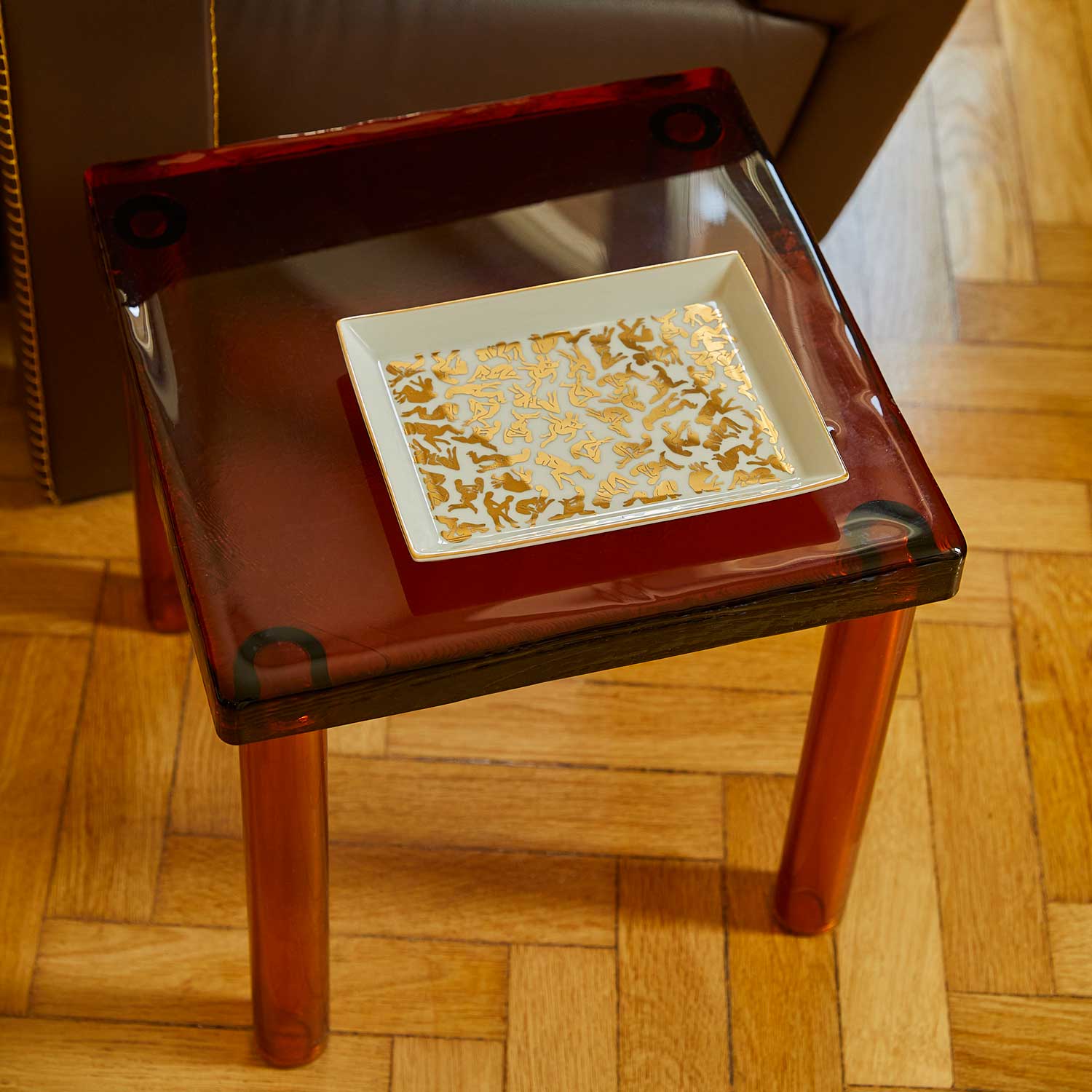 A white porcelain tray featuring a pattern of Kamasutra positions, resting on a small red glass table.