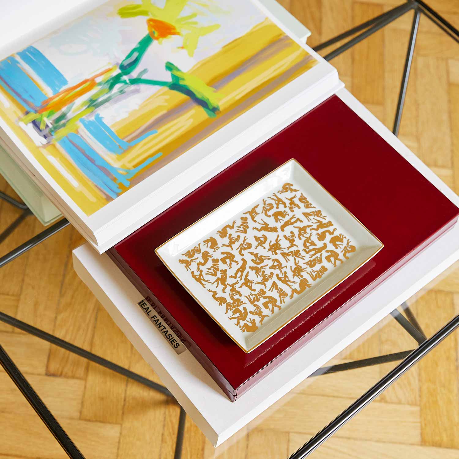 A white porcelain tray featuring a pattern of Kamasutra positions, along with lots of books and papers, on a small glass table.