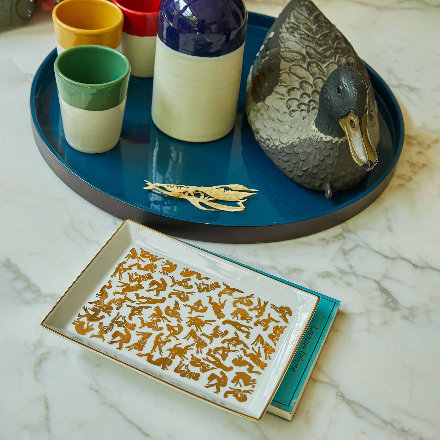 A white porcelain tray featuring a pattern of Kamasutra positions, with colourful ceramic glasses on a marble table.