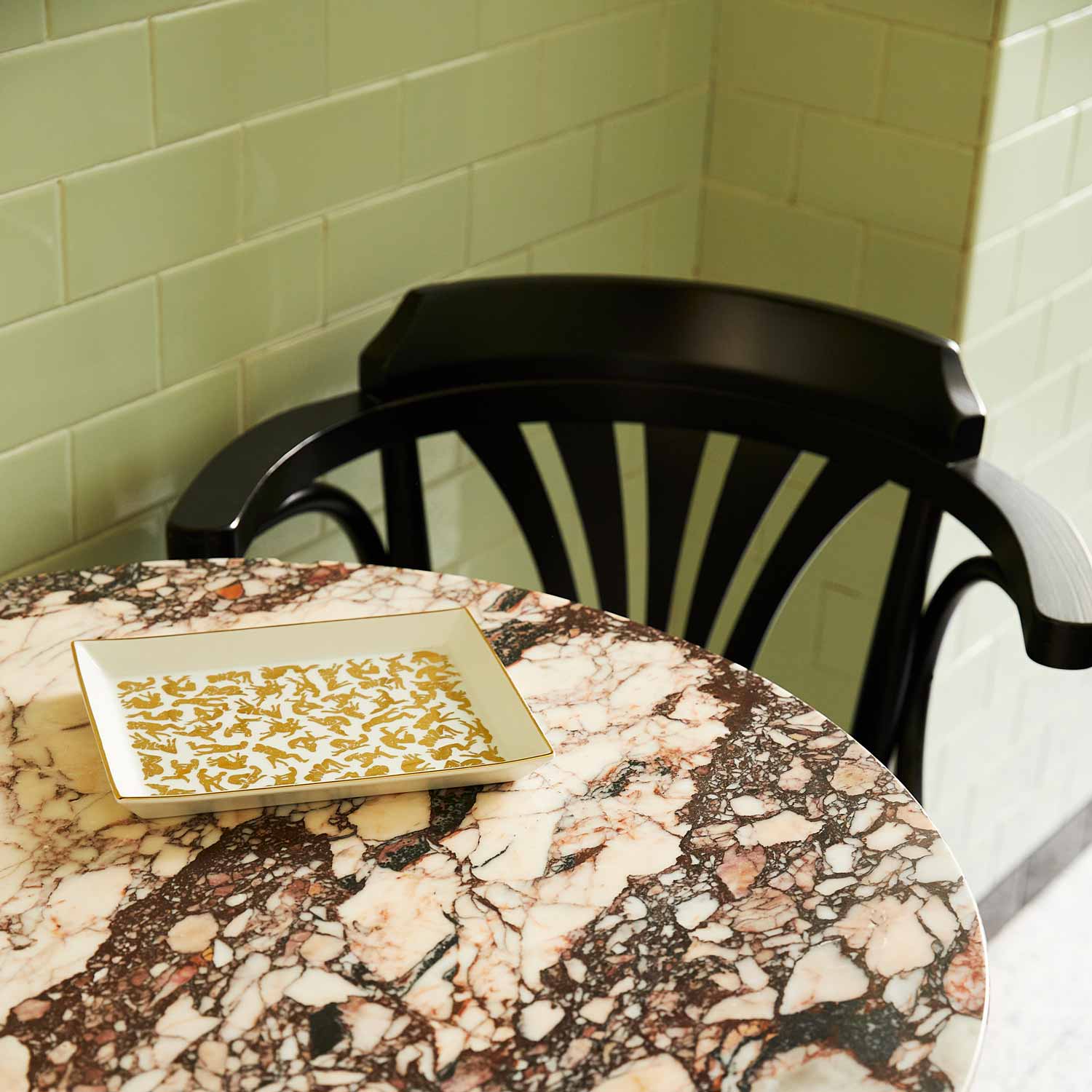 A white porcelain tray featuring a pattern of Kamasutra positions on a marble table next to a wooden chair.