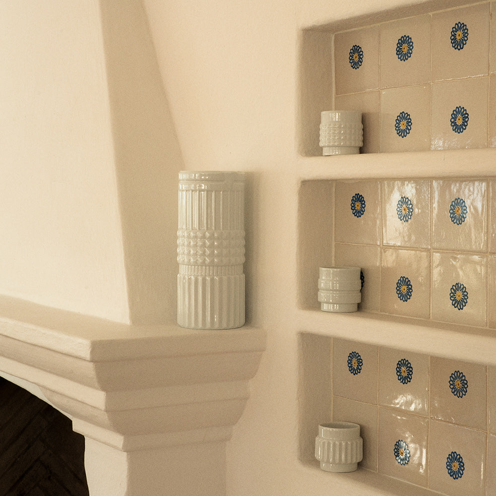 A white porcelain vase and glasses, whose shape is reminiscent of the various forms of nuts, sitting on shelves against a wall decorated with tiles.