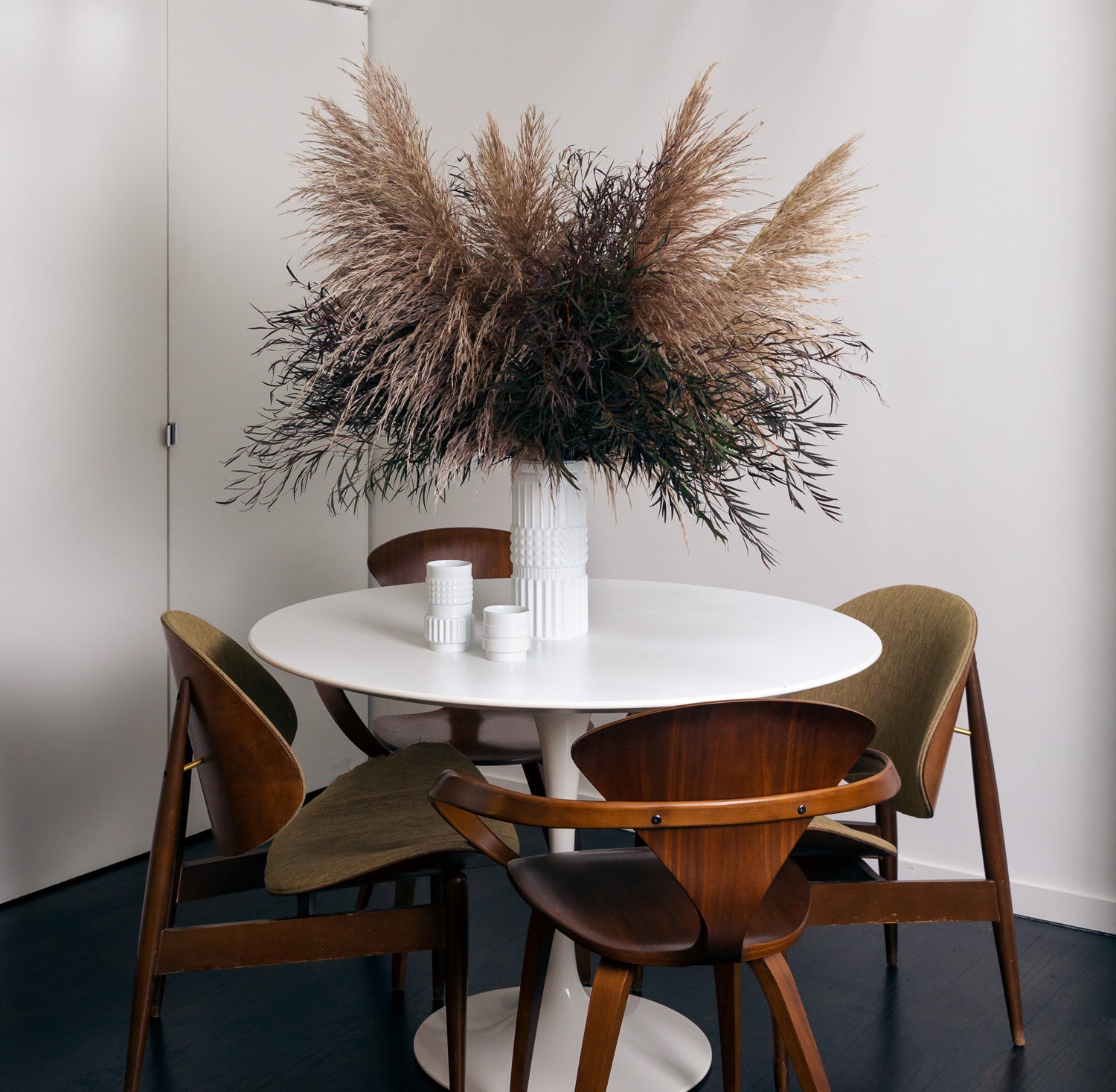 A white porcelain vase and glasses, whose shape is reminiscent of the various forms of nuts and bolts, sit on a designer table alongside four chairs. The vase contains dried flowers.