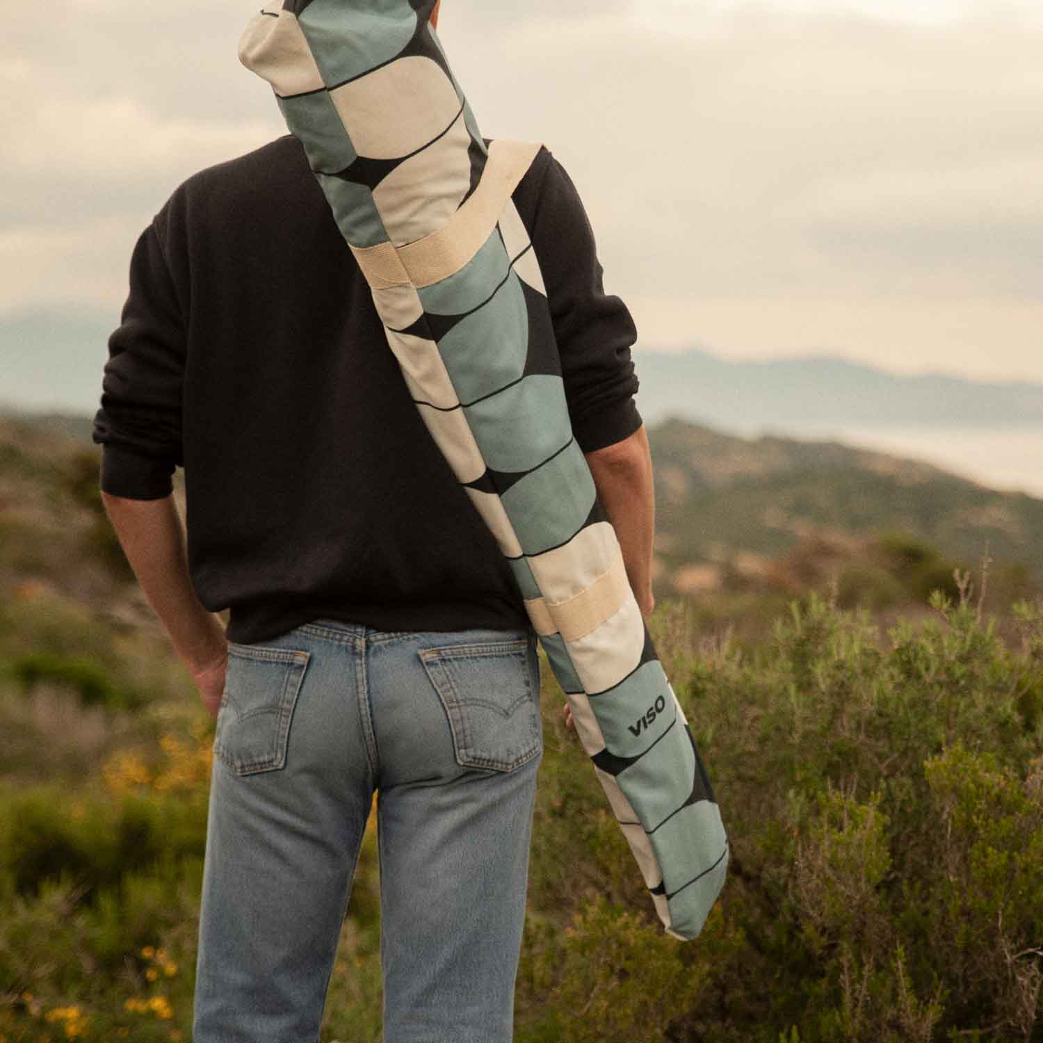 A boy seen from behind, carrying a bag containing a folded beach umbrella, looking out over a green field.