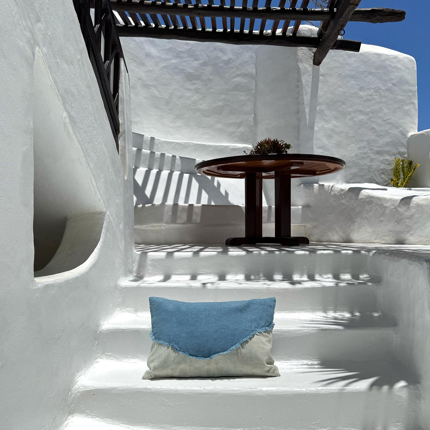 White-washed wall with a wooden bench, table, and blue pillow in a sunlit outdoor setting.
