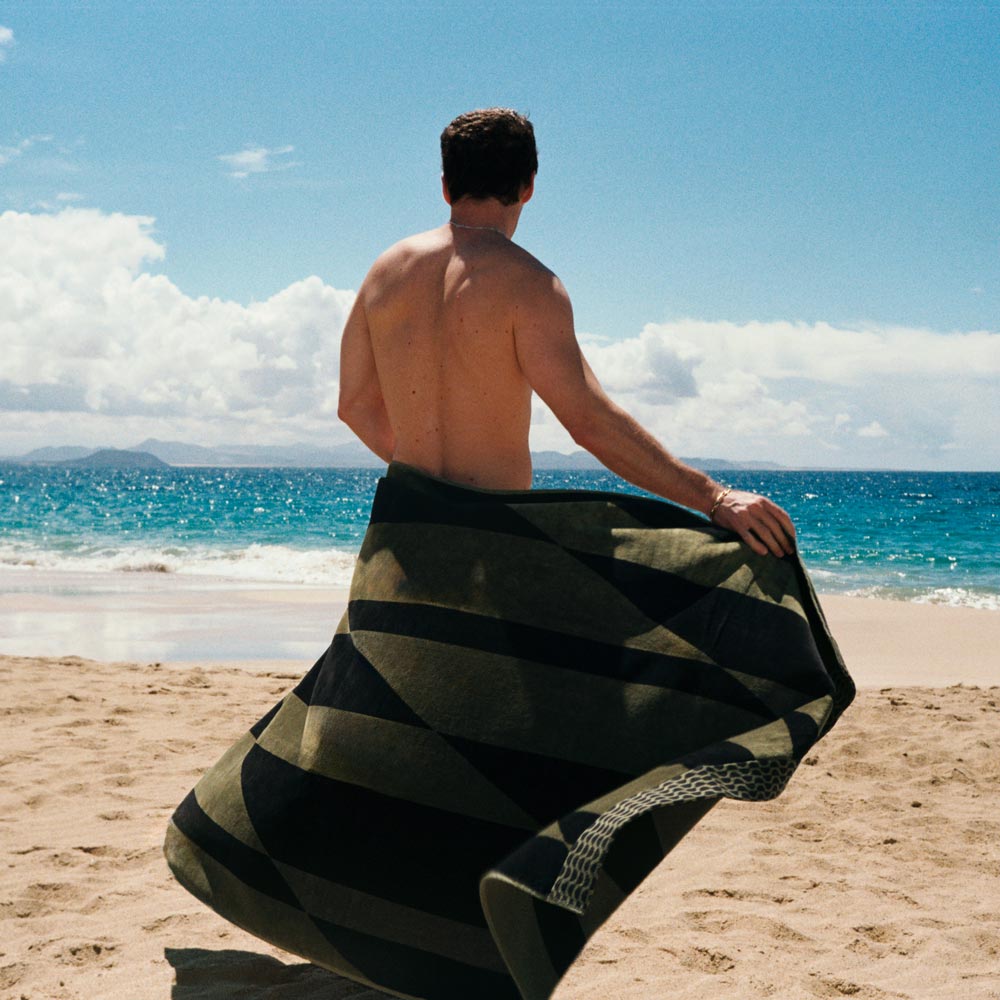 A boy with his back to the camera on a beach, wrapped in a green and black cotton towel.