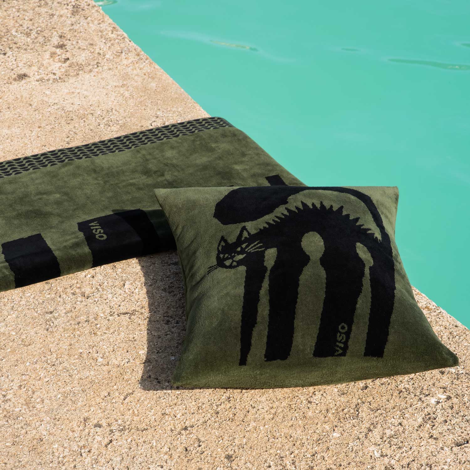 A green and black towel and pillow featuring a print of a cat by a swimming pool.