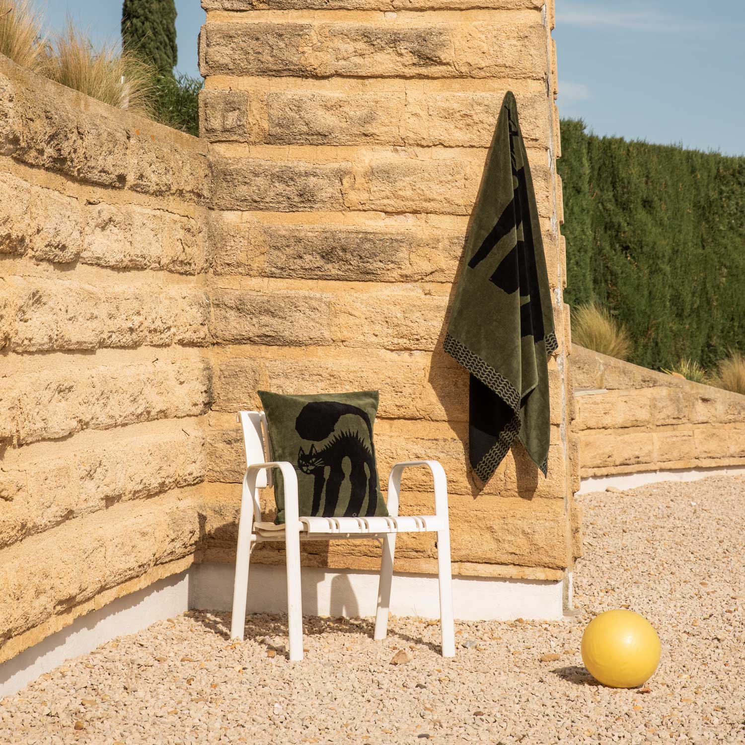 A towel hanging on the wall and a cushion on a white outdoor chair, both featuring a green and black cat print, alongside a beach ball.
