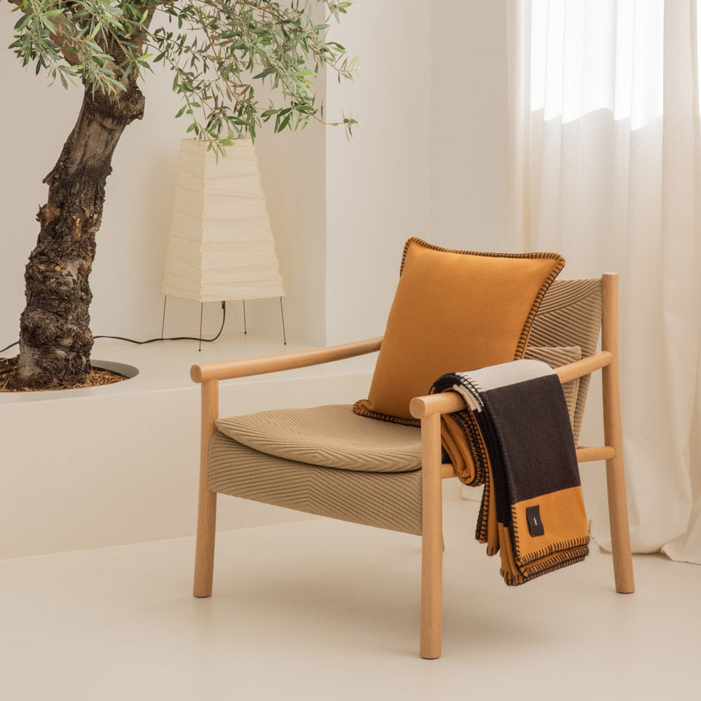 A living room with an armchair, on which there is a pillow and a merino blanket in black and camel, a lamp and a plant behind it.
