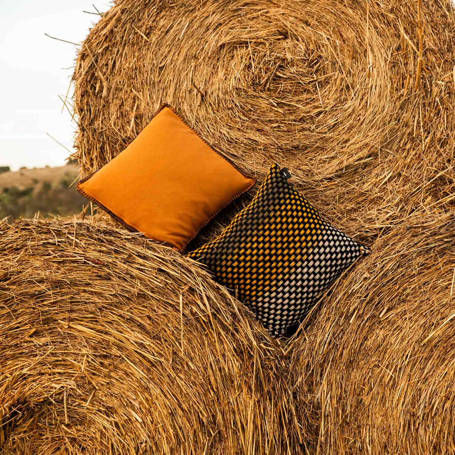 A brown merino pillow and a black, yellow and white basketweave wool pillow on some straw bales in the countryside.