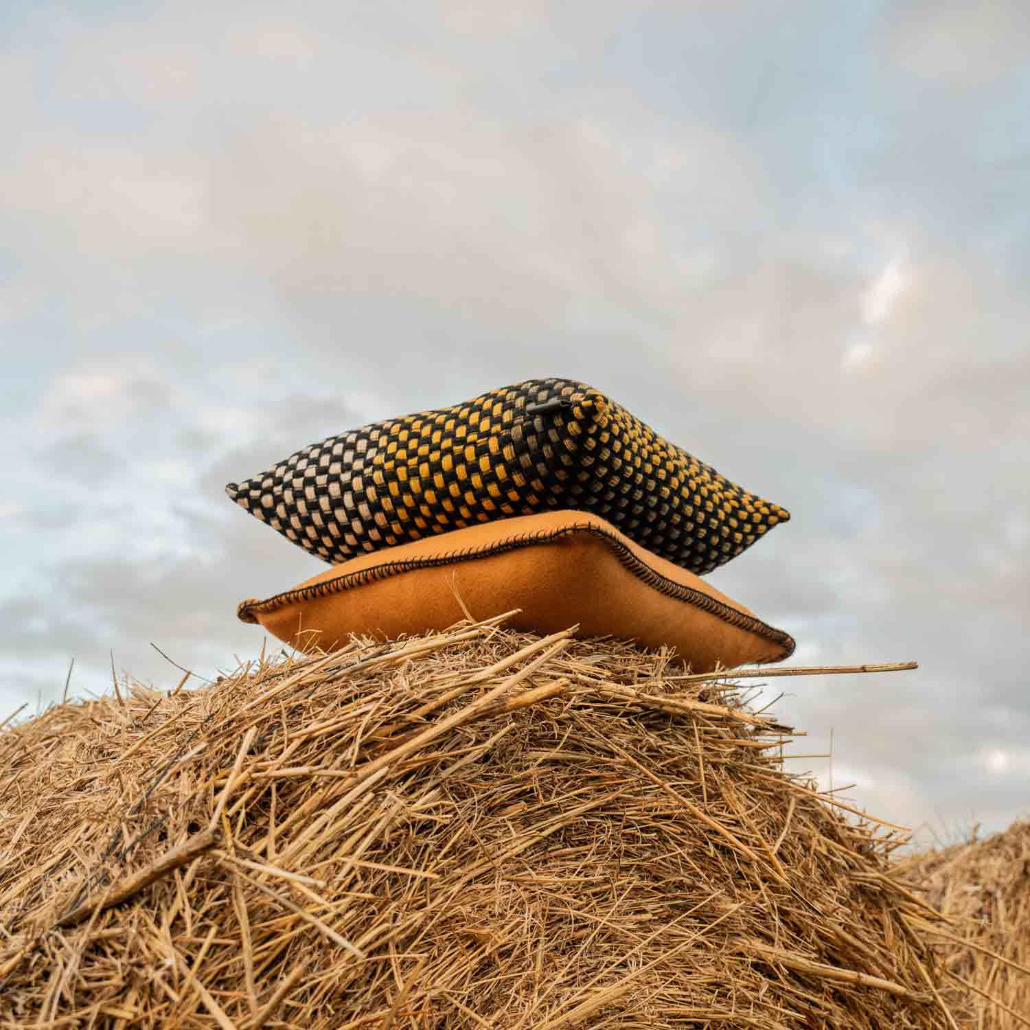A brown merino pillow and a black, yellow and white basketweave wool pillow on some straw bales in the countryside.