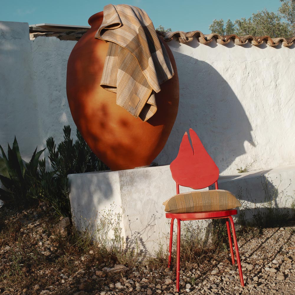 Red chair with a striped pillow next to a large brown pot against a white wall.