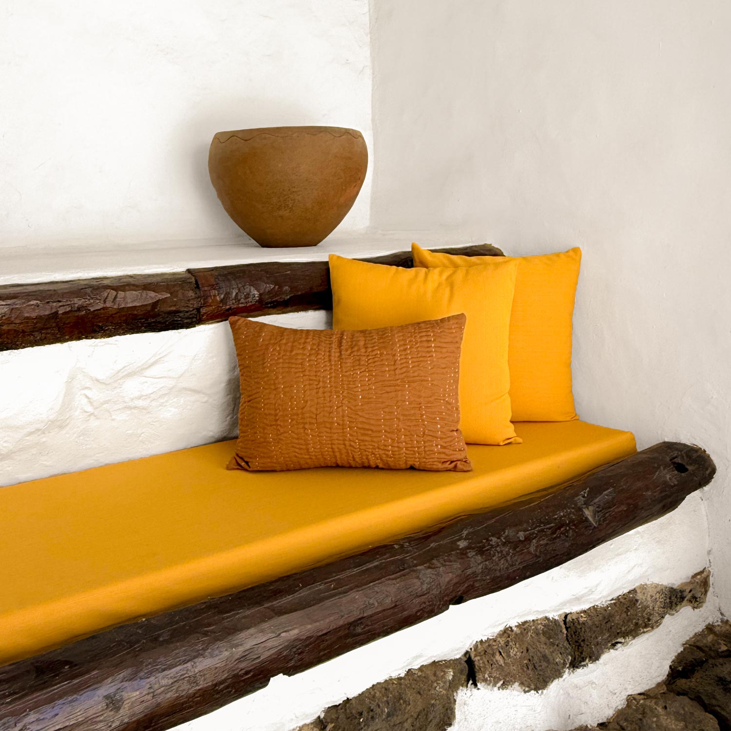 Wooden bench with yellow pilllows and a textured brown pillow against a white wall.