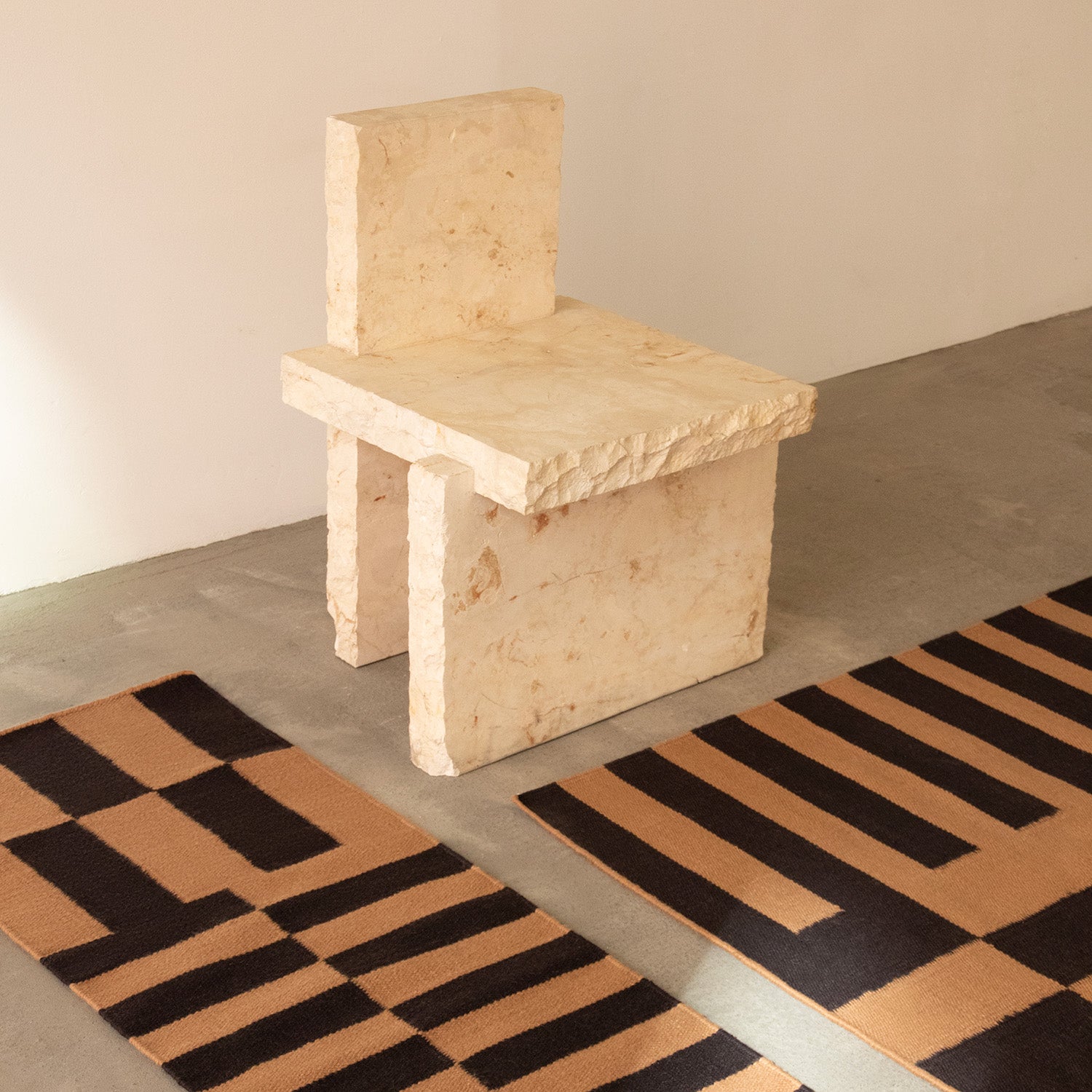 Beige stone chair on a concrete floor with geometric patterned rugs.